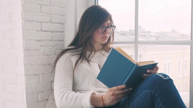 A young girl in glasses sits on the windowsill in a bright and cozy city room and reads a book. Slow motion.