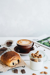 The cup of coffee, yoghurt with dried fruits and chocolate croissant at the white table.