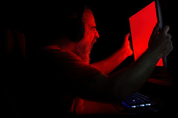 Angry bearded man in red light screaming in rage at PC monitor and shaking it violently  - obrazy, fototapety, plakaty