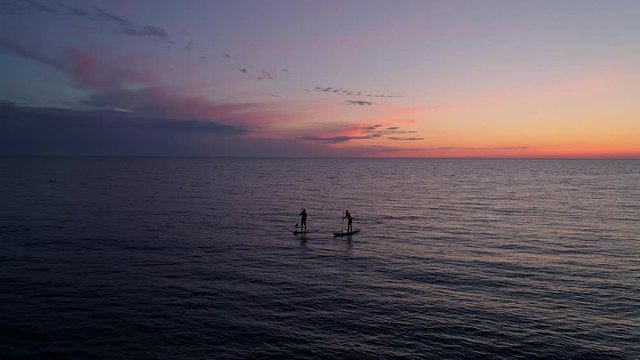 Aerial view couple silhouettes on paddle boards in sea during sunset. Amazing orange sky in background.