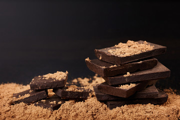 Broken dark chocolate and pile of cocoa on black background.