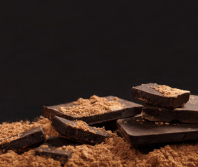 Broken dark chocolate and pile of cocoa on black background.