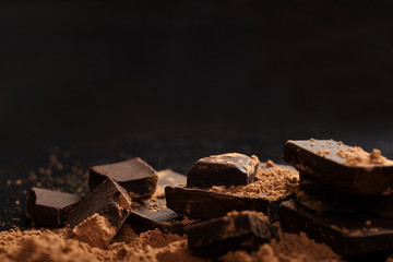 Broken dark chocolate and pile of cocoa on black background.