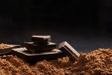 Broken dark chocolate and pile of cocoa on black background.