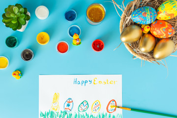 Easter concept, basket with hand made Easter eggs next to multicolor paints brush and white paper with sign happy Easter and paint multicolor Easter eggs on a blue bacground