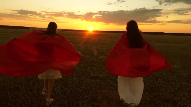 Mom and daughter play super heroes in red cloaks at sunset in the park. Girls play superheroes running across the field against the blue sky. concept of happy family. concept woman, savior, winner