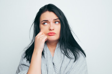 A young beautiful girl in a gray robe troraet his cheek, thinking about fat cheeks, about excess weight, and that it is time to lose weight on a gray-white studio background.