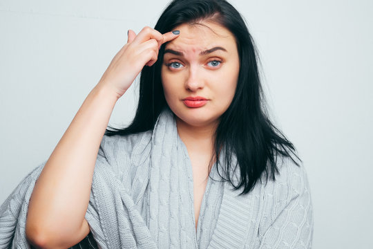 Upset girl in a gray robe indicates poor skin, age wrinkles, acne on a white and gray studio background. A woman is looking for flaws and age changes.