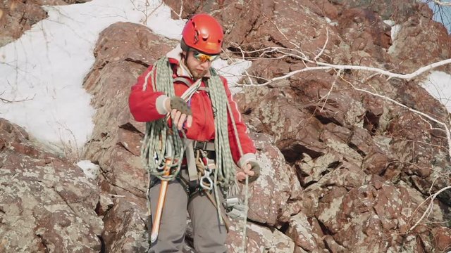 climber measures a long rope, throwing it on his shoulders, he is preparing for a dangerous descent. in the background, the rock is covered with snow and branches.