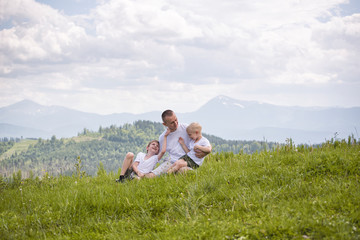 Naklejka na ściany i meble Happy father with his two young sons sitting on the grass on a background of green forest, mountains and sky with clouds. Friendship concept.