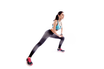 A dark-haired woman coach in a sporty  short top and gym leggings makes lunges  by the feet side, hands are held out to the side   on a  white isolated background in studio