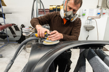 Professioneller Autolackierer schleift ein Autoteil mit der Schleifmaschine vor in einer Werkstatt - Serie Autowerkstatt - 259704566