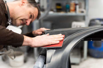 Professioneller Autolackierer schleift ein Autoteil mit Schmirgelpapier vor bei in einer Werkstatt - Serie Autowerkstatt.