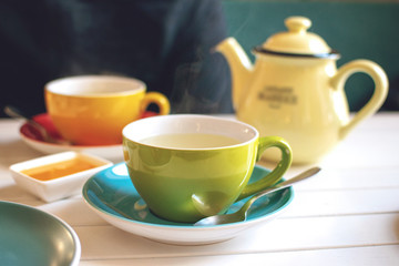 Herbal tea in green cup, honey and yellow teapot on white wooden table in cafe.  A cup of hot tea with steam. Natural light