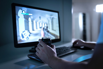 Gamer guy playing video game and drinking soda or energy drink from can. Fps videogame in computer monitor. Addicted gaming late at night. Young man with addiction to online first person shooters.