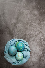 Easter painted eggs in a towel rolled up in a nest shape. Blue-green shades on a dark marble background. Easter holiday theme.