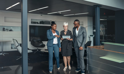 Three corporate business people walking out of a meeting discussing information on an digital tablet