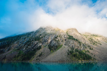 Naklejka na ściany i meble Giant cloud above rocky ridge with trees in sunlight. Amazing mountain lake. Mountain range under blue cloudy dawn sky. Wonderful rocks. Morning landscape of highland nature. Low clouds in sunny light