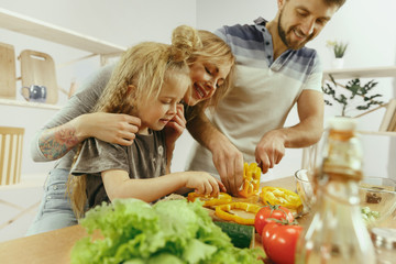 Cute little girl and her beautiful parents are cutting vegetables and smiling while making salad in kitchen at home. Family lifestyle concept