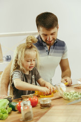 Cute little girl and her beautiful parents are cutting vegetables and smiling while making salad in kitchen at home. Family lifestyle concept