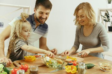 Cute little girl and her beautiful parents are cutting vegetables and smiling while making salad in kitchen at home. Family lifestyle concept