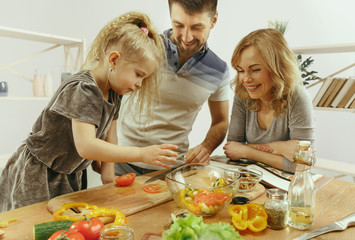 Cute little girl and her beautiful parents are cutting vegetables and smiling while making salad in kitchen at home. Family lifestyle concept