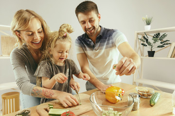 Obraz na płótnie Canvas Cute little girl and her beautiful parents are cutting vegetables and smiling while making salad in kitchen at home. Family lifestyle concept