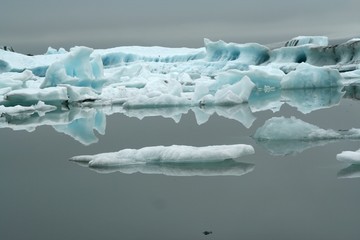 Reflections of crystal blue and white icerbergs in black dark water in dim gloomy light -...