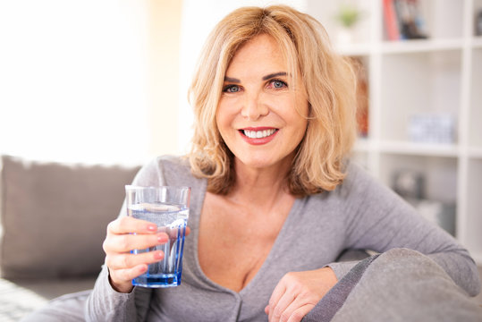 Best Ager, Frau nett lächelnd zuhause auf der Couch mit einem Glas Wasser 