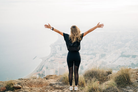 Sporty young woman enjoying freedom with open hands on the top of mountain looking the city from above