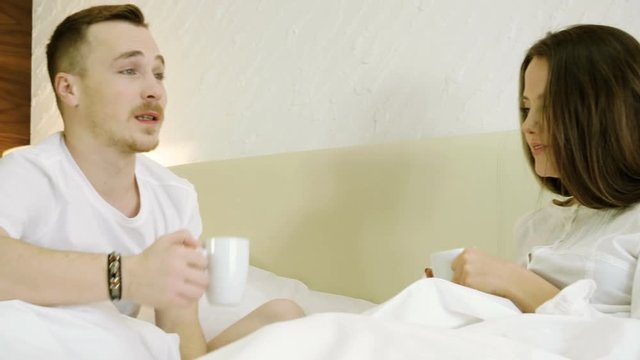 Young happy man and woman drinking coffee, talking, smiling and laughing in the bed. 4K