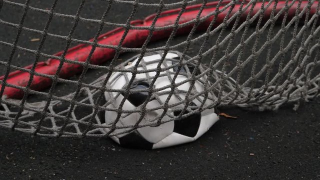 Broken deflated soccer ball lie in weary net of small goal gate, lost or unsuccessful game concept. Ball covered with net lie on black rubber surface of game of five-a-side field