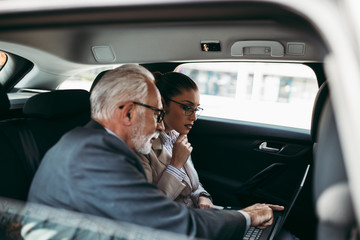 Good looking senior business man and his young woman colleague or coworker sitting on backseat in luxury car. They talking, smiling and using laptop and smart phones.