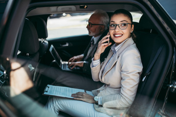 Good looking senior business man and his young woman colleague or coworker sitting on backseat in luxury car. They talking, smiling and using laptop and smart phones. 