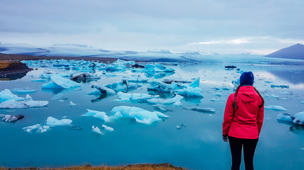 A young woman standing on the side of the glacier lagoon. Huge and massive ice bergs are slowly drifting towards the sea. Girl wears pink jacket. Pink contrasted with cold blue tones. Global warming © Chris