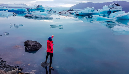 A girl in pink jacket standing on the rock in a shallow water. She is pointing at the drifting ice bergs in glacier lagoon. Glacier has many shades of blue. Global warming causing melting of glaciers © Chris