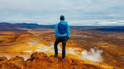 Young man wearing a blue jacket stands on top of a mud mountain, overlooking a geothermal spot noted for its bubbling pools of mud & steaming fumaroles emitting sulfuric gas. Power of planet Earth.