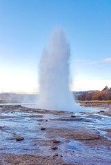 The Great Geysir, active geyser. Hot spring in a geothermal area. Steam of water bursts high up in the sky, splashing water all around. Water flowing on the rocks. Clear blue sky.