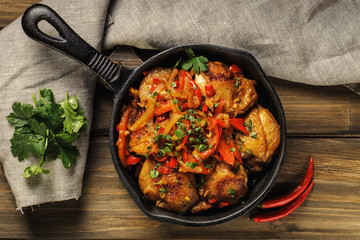 Fried chicken in spicy sauce with vegetables. Cooked in a cast iron pan. The pan is on a dark wooden background. Next is a natural linen napkin. Top view. Horizontal.
