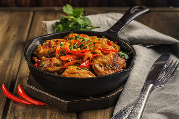 Fried chicken in spicy sauce with vegetables. Cooked in a cast iron pan. The pan is on a dark wooden background. Next is a natural linen napkin.
