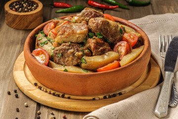 The traditional pork and potato dish of Georgian cuisine on earthenware ketsi. Ojahuri with tomatoes, hot peppers and fresh herbs. Dark background and natural napkin. Horizontal view.