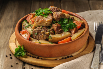 The traditional pork and potato dish of Georgian cuisine on earthenware ketsi. Ojahuri with tomatoes, hot peppers and fresh herbs. Dark background and natural napkin. Horizontal view.