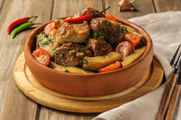 The traditional pork and potato dish of Georgian cuisine on earthenware ketsi. Ojahuri with tomatoes, hot peppers and fresh herbs. Dark background and natural napkin. Horizontal view.