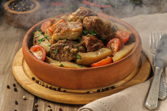 The traditional pork and potato dish of Georgian cuisine on earthenware ketsi. Hot Steam Dish. Ojahuri with tomatoes, hot peppers and fresh herbs. Dark background and natural napkin. Horizontal view.