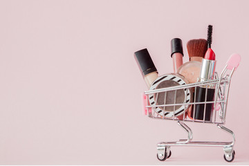 Creative concept with shopping trolley with makeup on a pink background. Perfume, sponge, brush,...