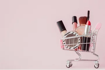 Poster Creative concept with shopping trolley with makeup on a pink background. Perfume, sponge, brush, mascara, pencil, nail file, eye shadow, lip gloss in the basket, copy space © volody10