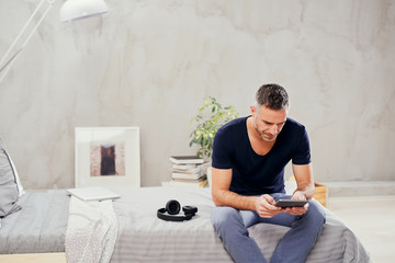 Caucasian man in forties sitting on bed in bedroom and using tablet.