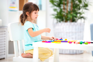 A pretty girl of three years old sits at the table and sculpts figures from colored clay. Sculpting and children's creativity