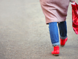 Woman in blue jeans, red boots, elongated coat with a package in hand go on the road. Rear view, blurred background, lifestyle