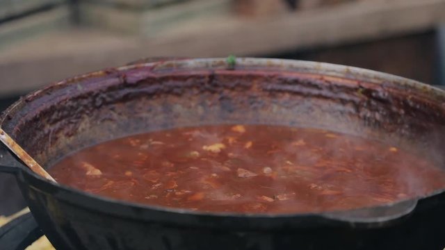 Close up slow motion of big iron cast kettle pot with a kitchen metal spoon with roast of meat boiling in tomato sauce. Street food roast pieces of meat boiling in big kettle in open space